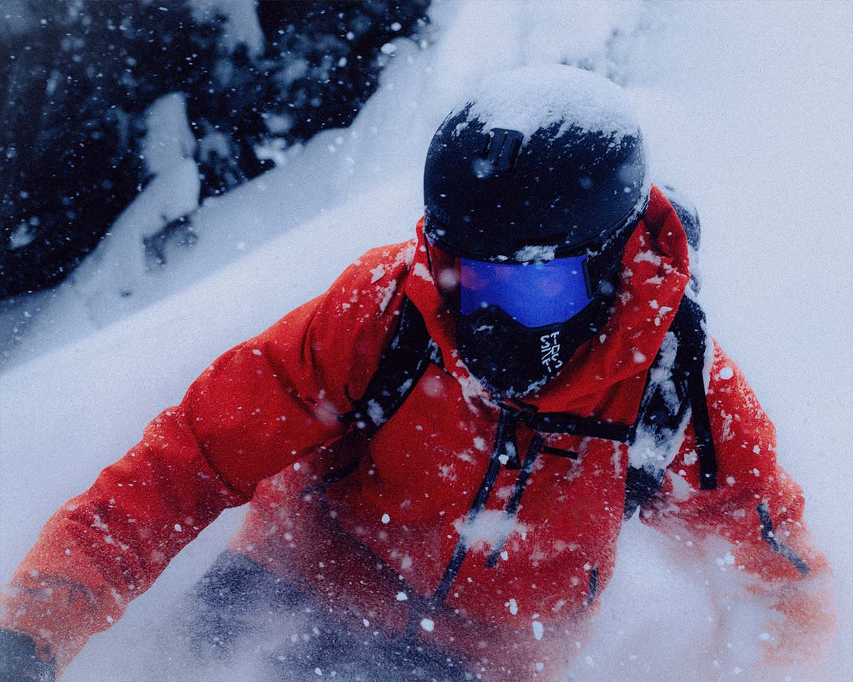 A skier in a red jacket and a STRATUS balaclava, turning through deep powder