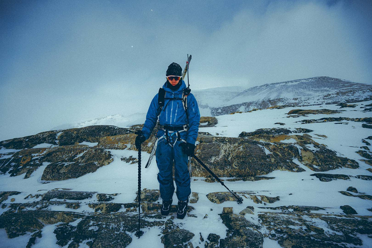 A ski mountaineer in blue on rocky, snow-covered ground, skis on the pack