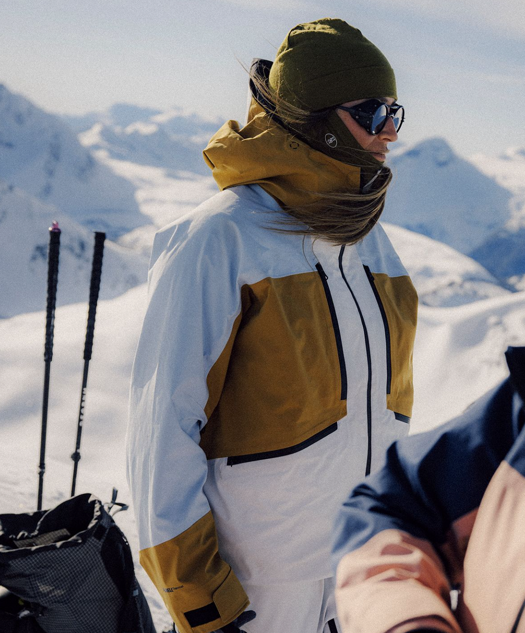 Two people in winter gear standing on a snowy mountain with skis and backpacks.
