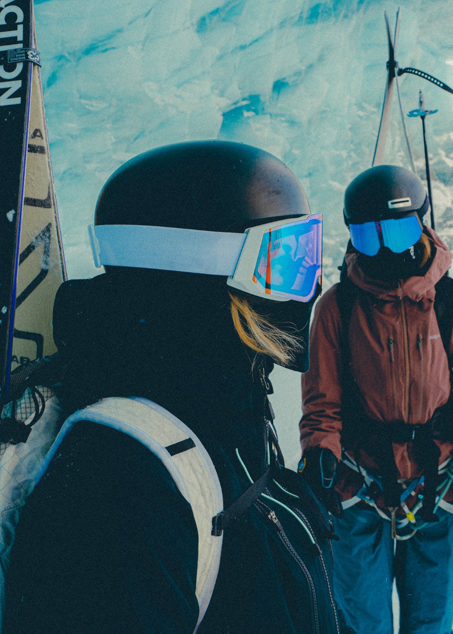 Two skiers with helmets and goggles standing against a snowy mountain backdrop.