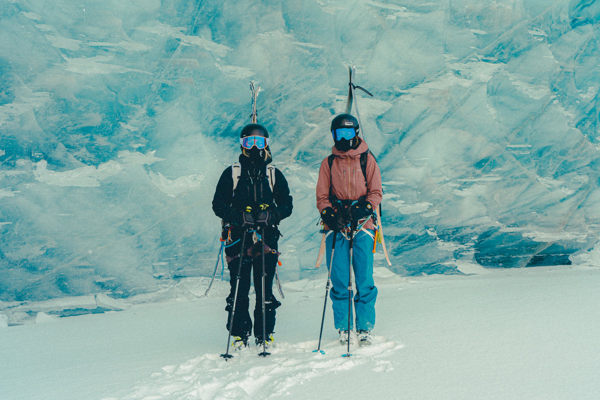 Two skiers in helmets and goggles stand in front of a blue glacier ice wall