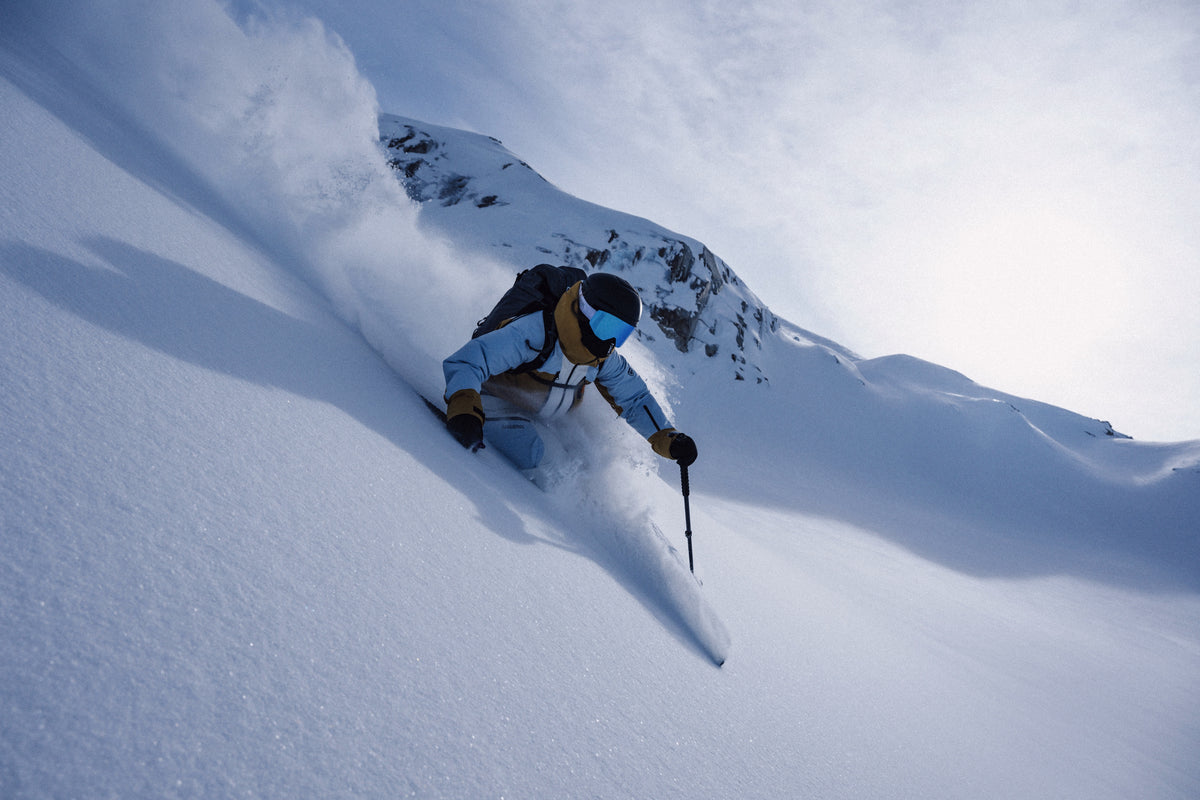 A skier in a light blue jacket and goggles carving through deep powder on a steep slope
