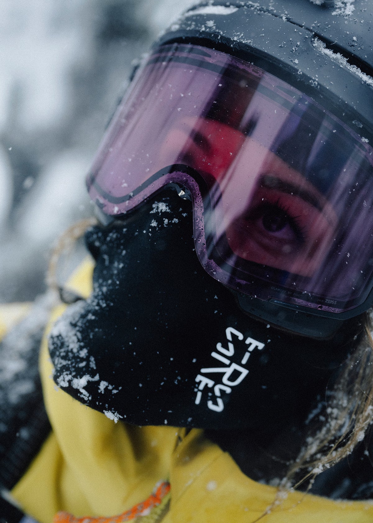 Close-up of a skier in pink-tinted goggles and a black STRATUS mask in falling snow