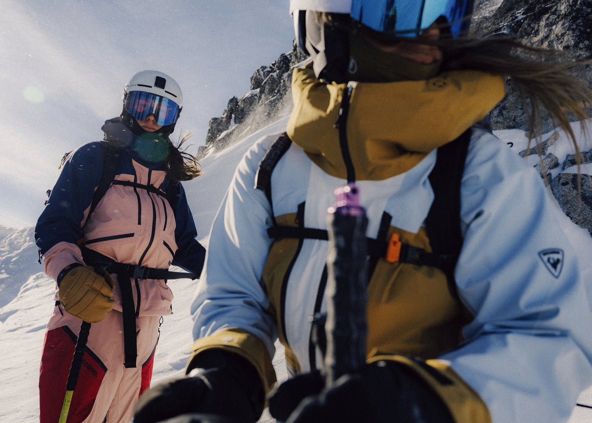 Two skiers in goggles and helmets on a snowy ridge, one close to the camera